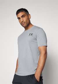 Sivi pamučni majica, kratki rukavi, okrugli izrez. Ima crni Under Armour logo na lijevoj strani prsnog koša. Glatka tekstura, ležeran kroj.