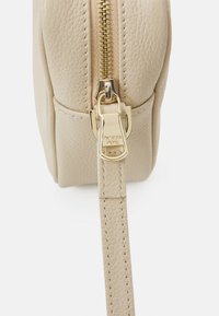 Borsa a mano in pelle beige con finitura texturizzata, caratterizzata da una cerniera e accessori in tonalità oro, e una tracolla piatta attaccata per il trasporto.