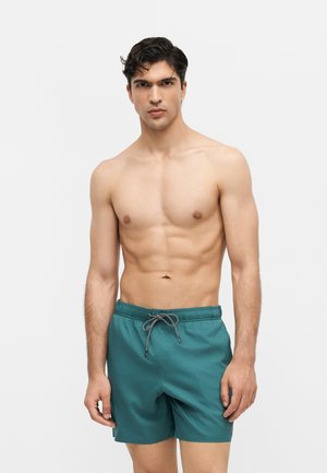 Młody mężczyzna o ciemnych włosach, noszący turkusowe kąpielówki ze sznurkiem, stojący na tle jednolitego jasnego tła, zwrócony przodem.