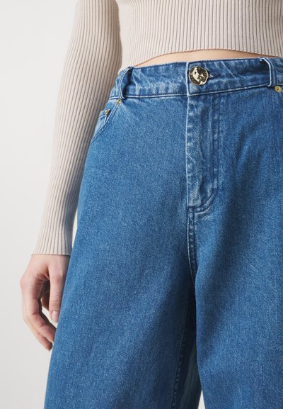 Un jean en denim bleu clair avec une taille haute, une fermeture à bouton doré et des détails de couture subtils. Complété par un haut beige côtelé.