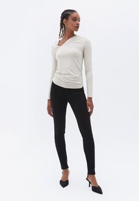 Top beige de manga larga con un escote asimétrico drapeado y detalle fruncido, combinado con pantalones ajustados negros y tacones negros de punta.