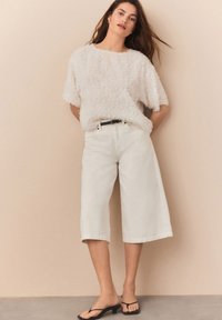 Maglione bianco dalla texture fluffy abbinato a culotte bianche a vita alta. Cintura nera che mette in risalto la vita con flip-flops neri.