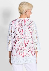 Haut en dentelle blanche avec un design découpé floral rose, manches trois-quarts et coupe ample, doté d'un col arrondi et de motifs détaillés.