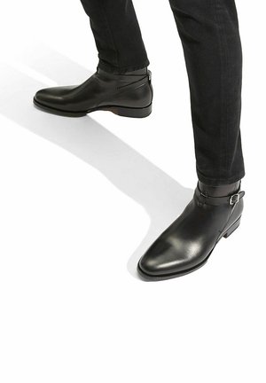 Botas de cuero negro hasta el tobillo con punta afilada y detalle de hebilla, que presentan una textura suave y un tacón bajo apilado.