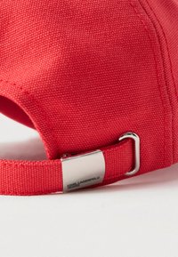 Alça de boné de beisebol em tecido vermelho com fivela de metal prateado gravada com "KARL LAGERFELD JEANS" sobre um fundo branco.