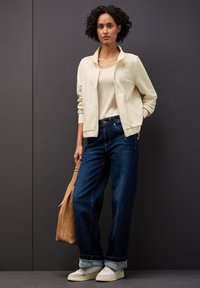 Giacca bomber beige chiaro, top crema, jeans a gamba larga blu scuro e sneaker bianche, abbinati a una borsa in suede color cammello. Sfondo grigio.
