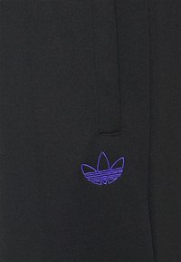 Tessuto nero con un piccolo logo ricamato adidas Trefoil di colore viola vicino a una cucitura verticale o all'apertura di una tasca.