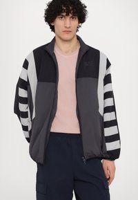 Vindjakke i sort, grå og hvid. Har striped ærmer, lynlås foran og et EA7-logo på brystet. Letvægts, tekstureret stof.