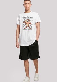 Hvit bomull t-skjorte med et grafisk trykk av en Tasmanian Devil, kombinert med svarte shorts og hvite joggesko. Enkel rund hals og avslappet passform.