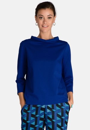 Blaues Langarmshirt mit einem Schalkragen und einer glatten Textur. Kombiniert mit gemusterten Hosen, die geometrische Formen in Blau und Schwarz aufweisen.