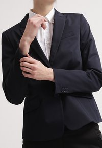 Persona vestida con un blazer azul marino oscuro y una camisa blanca de botones, ajustando el cuello del blazer con los brazos cruzados.