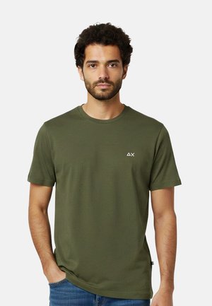 Uomo con capelli corti e ricci e barba, che indossa una t-shirt a maniche corte verde oliva con un piccolo logo bianco sul petto, in piedi con una mano in tasca.