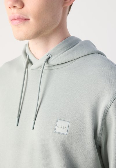 Gros-plan sur une personne portant un sweat à capuche gris clair avec des cordons de serrage et un petit logo carrée "BOSS" sur la poitrine.