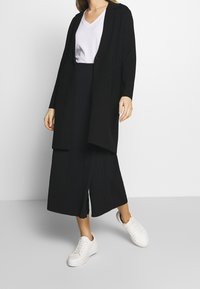 Svart lång cardigan med avslappnad passform, bärs över en vit V-ringad t-shirt och svarta vida byxor, ihop med vita sneakers.