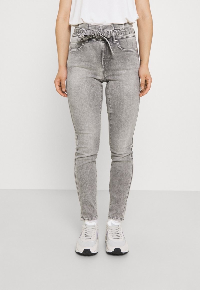 Jeans grises de tiro alto con cinturón anudado, ajuste slim y bolsillos frontales. Llevados con zapatillas blancas sobre un fondo liso.