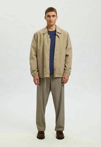 Világos bézs színű dzseki patentos gombokkal, rugalmas mandzsettával, sötétkék ing fölött viselve. Bő szürke nadrág, barna cipő. Semleges háttér.