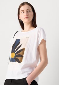 T-shirt à manches courtes blanche avec un imprimé graphique d'une fleur aux feuilles bleues et noires et un cercle jaune sur un fond beige.