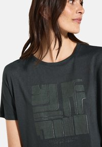 Camiseta gris oscuro con un estampado texturizado y geométrico en verde que presenta formas abstractas y el texto "CREAR PARA INSPIRAR" en la parte inferior.