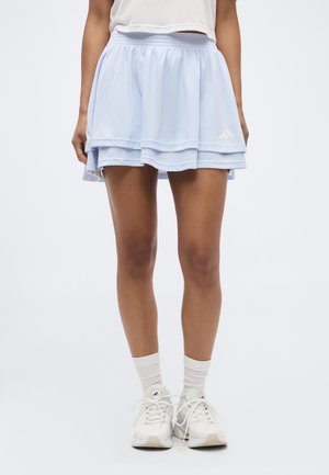 Nohy osoby oblečenej v svetlomodrej vrstvenej tenisovej sukni, bielych ponožkách a bielych teniskách Adidas stojacej na bielom pozadí.