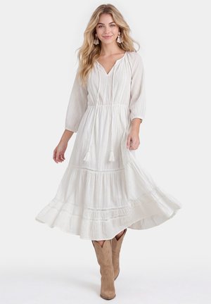 Vrouw die een lange witte jurk draagt met driekwartmouwen en beige cowboylaarzen, staand tegen een effen lichte achtergrond.