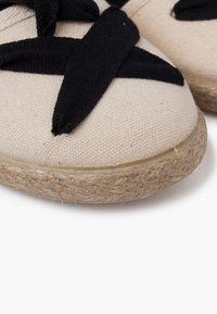 Espadrillas in tela beige con lacci in tessuto nero, dotate di suola in juta intrecciata e bordo in gomma liscia. Superficie testurizzata con cuciture visibili.