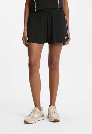 Kvinde iført sorte atletikshorts med hvidt logo på venstre ben og beige sneakers, stående mod en ensfarvet hvid baggrund.