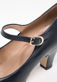 Chaussure Mary Jane en cuir noir avec talon bloc de hauteur moyenne et bride à boucle dorée sur doublure intérieure beige.