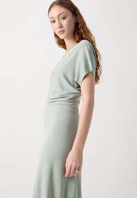 Svijetlozelena haljina do koljena od mekog, glatkog materijala. Ima opušteni kroj s V-izrezom i kratkim rukavima. Jednostavan i elegantan dizajn.