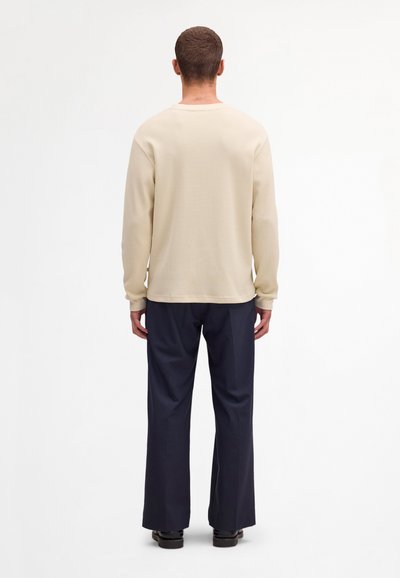 Homme de dos portant une chemise crème texturée à manches longues, un pantalon large marine et des chaussures noires sur fond blanc.