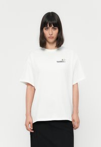 Mujer con el cabello oscuro y lacio, vistiendo una camiseta blanca de gran tamaño con un pequeño abejorro y el texto "Converse Thank You For Growing" en el pecho.
