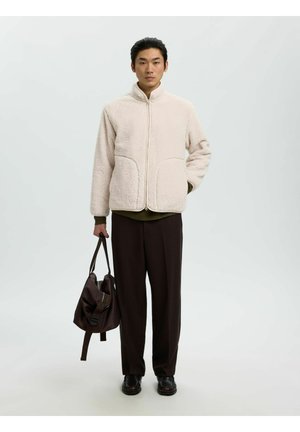 Homme portant une veste polaire beige et un pantalon noir tenant un sac marron foncé dans une main sur un fond clair uni.