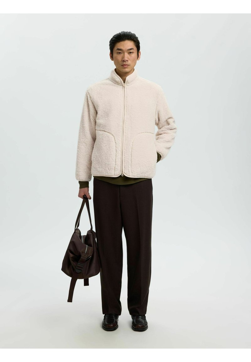 Homme portant une veste polaire beige et un pantalon noir tenant un sac marron foncé dans une main sur un fond clair uni.