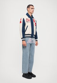 Varsityjakke i creme og marineblå med broderede applikationer; afslappede denimjeans med pink katte-design; sorte sko.