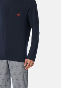Marineblauw shirt met lange mouwen en een klein rood logo op de borst, gecombineerd met grijze pyjamabroek met een boompatroon. Zachte stof, casual ontwerp.
