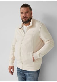 Homme barbu avec les cheveux attachés, portant une veste beige zippée sur une chemise blanche, se tenant avec une main dans sa poche.