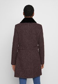 Cappotto tessuto bordeaux con colletto in pelliccia sintetica nera, vita cinturata e maniche lunghe; presenta un motivo testurizzato su tutta la superficie.