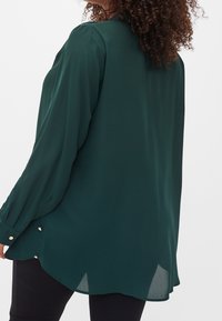 Blusa fluida di un verde scuro, con maniche lunghe, dettaglio dei bottoni sui polsini e orlo curvo, realizzata in tessuto leggero.