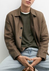 Homme portant une veste marron à boutons par-dessus un pull vert foncé et un jean bleu clair, assis avec les mains lâchement croisées sur les genoux.