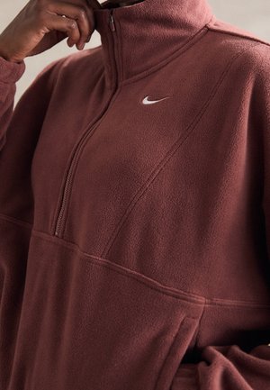 Osoba nosząca brązową bluzę polarową z zamkiem do ćwiartki, trzymająca zamek w pobliżu szyi, z małym logo Nike.