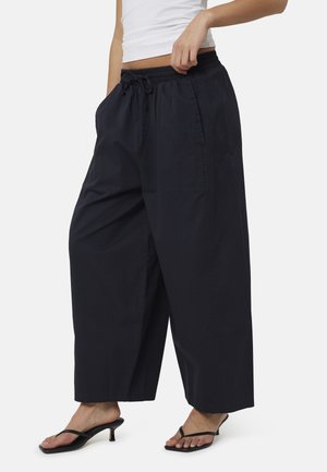 Persoon draagt losse zwarte wijde broek met trekkoord in de taille en zwarte sandalen met lage hak, houdt de taille met één hand vast.