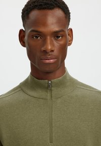 Jeune homme aux cheveux courts bouclés portant un pull zippé vert olive regardant droit devant sur un fond uni.