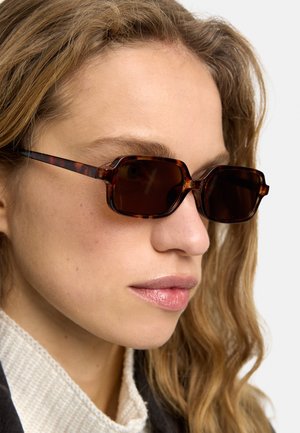 Młoda kobieta o falowanych blond włosach, nosząca prostokątne okulary przeciwsłoneczne w oprawce typu „żółw” oraz biały dziergany sweter.