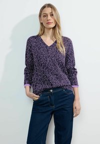 Top viola a scollatura a V con un motivo testurizzato in tonalità di viola più chiaro, dotato di maniche lunghe con polsini color lavanda. Abbinato a jeans blu scuro.