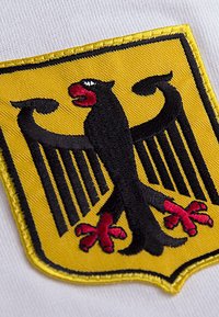 Black eagle with red beak and claws on a yellow shield-shaped patch sewn onto white fabric.