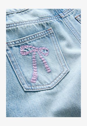 Lyseblåt denimstof med en syet lilla sløjfe på baglommen. Teksturen er glat med synlige syningsdetaljer.