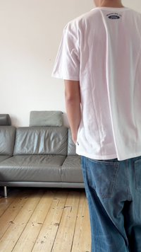 Baltas medvilninis marškinėlis su mažu logotipu nugaroje, derinamas su laisvo stiliaus mėlynais džinsais. Aplinkoje matomas pilkos odos sofa.