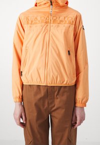 Jasnopomarańczowa kurtka zapinana na zamek z kapturem, z frontowym nadrukiem logo, elastycznymi mankietami i jedną boczną kieszenią. Noszona na brązowych spodniach.