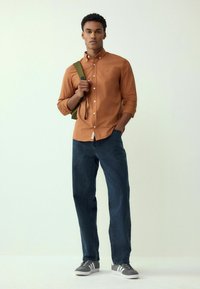 Pruun nuppudega särk, tume teksapüksid, roheline seljakott, hallid tennisetossud valgete aktsentidega. Mudel seisab heleda tausta vastu.