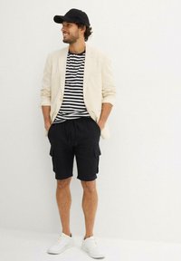 Blazer beige sopra una maglietta a righe bianco e nera, pantaloni cargo neri e sneakers bianche. Un cappellino nero completa il look informale.