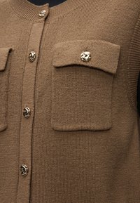 Καφέ πλεκτό γιλέκο με κουμπώματα μπροστά και δύο τσέπες στο στήθος με καπάκι, η κάθε μία ασφαλισμένη με ένα λαμπερό χρυσό διακοσμητικό κουμπί.
